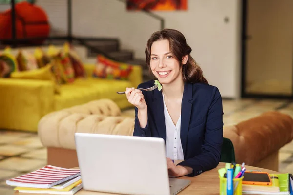 Jovem feliz no escritório no laptop com garfo com salada — Fotografia de Stock