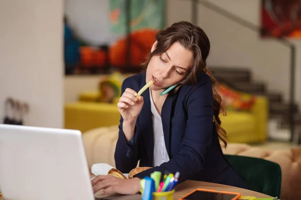 Mujer en el lugar de trabajo con teléfono inteligente portátil, comer papas fritas . — Foto de Stock