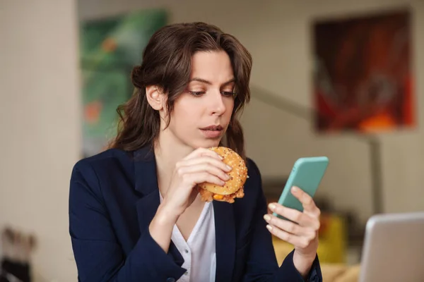 Mujer joven interesada seria con hamburguesa y teléfono inteligente — Foto de Stock