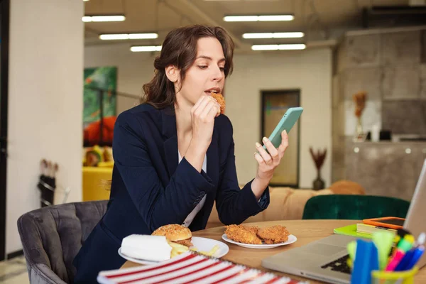 Mujer comiendo hamburguesa sentado en la oficina moderna en el lugar de trabajo . — Foto de Stock