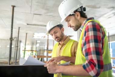 Sarı yelekli inşaat işçileri dikkatle planlar üzerinde çalışıyorlar.