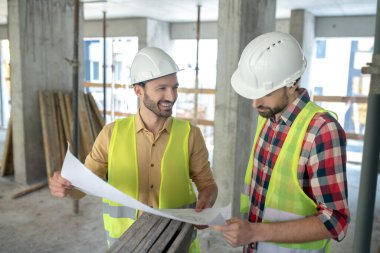 Sarı yelekli, gülümseyen bina işçisi elinde planı meslektaşına gösteriyor.