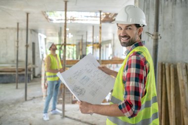Sarı yelekli bir bina işçisi kafasını kameraya çeviriyor, elinde planlar, arka planda duran meslektaşı.