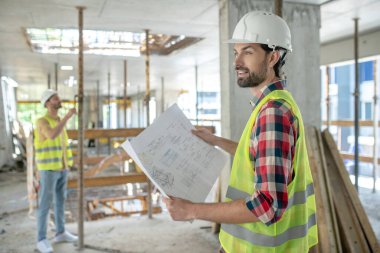 Sarı yelekli bir bina işçisi elinde planlar, arka planda duran meslektaşı, inşaatı kontrol ediyor.