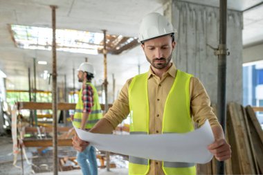 Sarı yelek giymiş bir bina işçisi planlar üzerinde çalışıyor. İş arkadaşı arka planda duruyor, inşaatı kontrol ediyor.