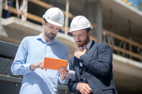 Foreman ukazuje něco na svém tabletu vedoucímu budovy, který se dotýká jeho brady — Stock fotografie