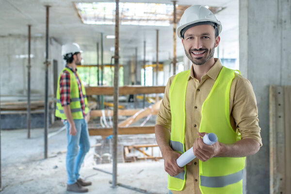 Building worker in yellow vest holding rolled blueprint, smiling, his colleague standing in the background