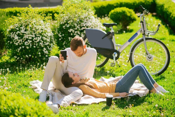 Mężczyzna siedzący z kawą, kobieta leżąca na plecach na trawie — Zdjęcie stockowe