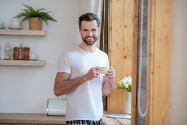 Koyu renk saçlı yakışıklı bir adam elinde kupa ve gülümsüyor.