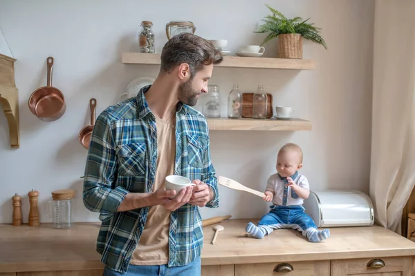 Junger Vater steht mit einer Tasse in der Hand und schaut seinen kleinen Sohn an — Stockfoto