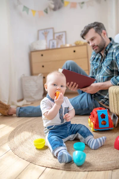 Apa kockás ingben, kezében egy könyvvel, a kisfia játékokkal játszik. — Stock Fotó
