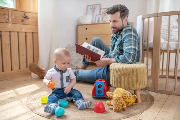 Padre con una camisa a cuadros sentado en el suelo con un libro, su pequeño hijo jugando con juguetes —  Fotos de Stock