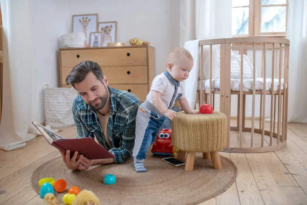 Vater im karierten Hemd liegt auf dem Boden und liest ein Buch, sein kleiner Sohn spielt neben ihm — Stockfoto
