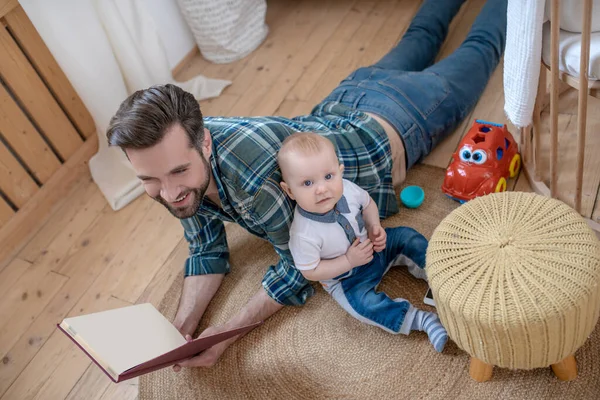 Pai em uma camisa quadriculada segurando um livro e seu filho sentado ao lado de hime no chão — Fotografia de Stock