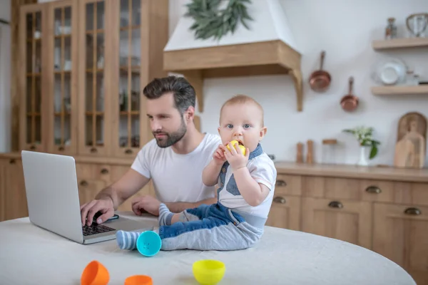 Dunkelhaariger Vater arbeitet, während sein kleiner Junge neben ihm auf dem Tisch sitzt — Stockfoto