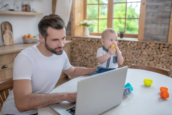 Ciemnowłosy ojciec pracujący nad laptopem, jego synek siedzący obok niego — Zdjęcie stockowe