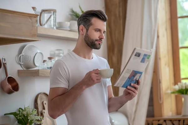 Homem bonito de cabelos escuros tomando café e lendo um jornal — Fotografia de Stock