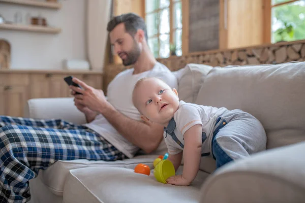 Leuke baby spelen op de bank, zijn vader kijken naar iets op een smartphone — Stockfoto