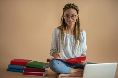 Genç Afro-Amerikalı kadın gözlük takıyor, dizüstü bilgisayarıyla yerde oturuyor, notlar alıyor, gülümsüyor.