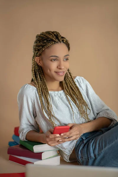 Joven mujer afroamericana sentada en el suelo, apoyada en un montón de libros, sosteniendo su móvil — Foto de Stock