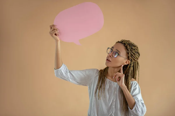 Giovane donna afroamericana in occhiali che tiene una nuvola di dialogo rosa, toccandosi il mento con l'indice — Foto Stock