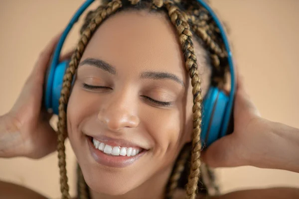 Sorrindo jovem afro-americano feminino em fones de ouvido azuis, ouvindo música com olhos fechados — Fotografia de Stock