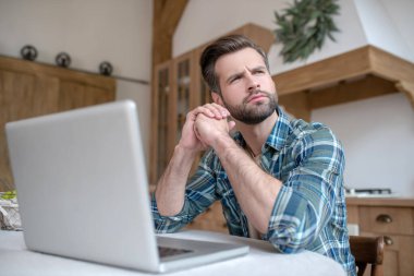 Kareli tişörtlü bir adam dizüstü bilgisayarın başında oturup düşünüyor.