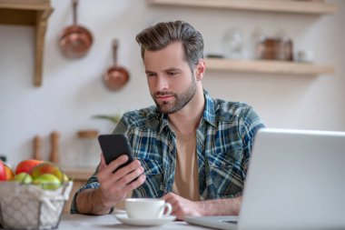 Kareli tişörtlü bir adam akıllı telefon tutuyor ve bir mesaj okuyor.