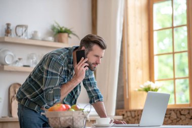 Kareli gömlekli bir adam telefonda konuşuyor ve meşgul görünüyor.