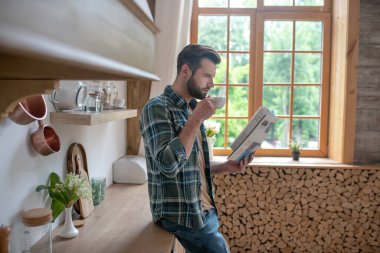 Kareli gömlekli, kahve içen ve gazete okuyan uzun bir adam.