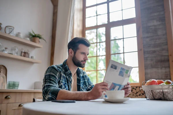 Hombre barbudo con una camisa a cuadros sentado en la mesa, leyendo el periódico buscando concentrado —  Fotos de Stock