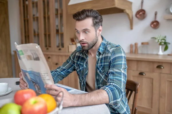 Muž v kostkované košili sedí u stolu, čte noviny a vypadá překvapeně — Stock fotografie