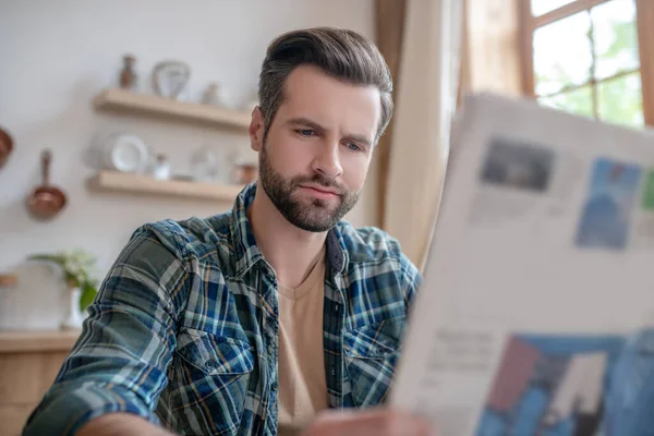 Muž v kostkované košili sedí u stolu, čte noviny a vypadá vážně — Stock fotografie