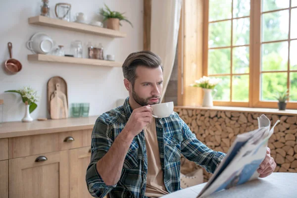 Hombre alto en una camisa a cuadros sentado a la mesa, tomando café y leyendo un periódico —  Fotos de Stock