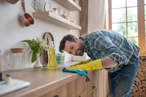 Темноволосый мужчина в клетчатой рубашке и в жёлтых перчатках чистит стол на кухне. — стоковое фото