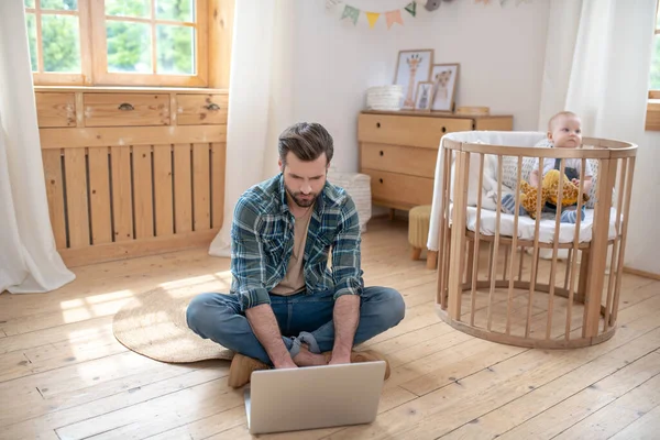 Donkerharige vader ziet er druk uit, werkt aan een laptop terwijl zijn zoontje speelt — Stockfoto