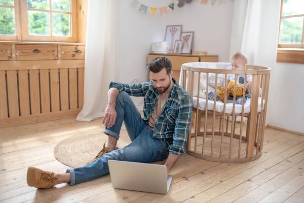 Donkerharige vader die op de vloer zit, werkend op een laptop terwijl zijn zoontje speelt — Stockfoto