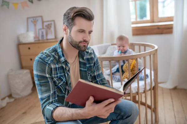 Ciemnowłosy ojciec czyta książkę, podczas gdy jego mały syn bawi się — Zdjęcie stockowe