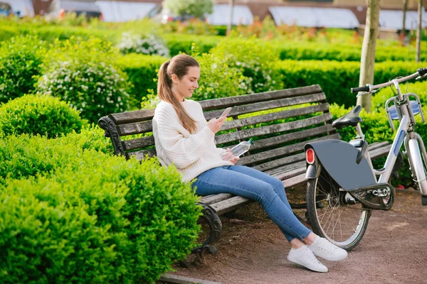 Dziewczyna patrząc w smartfon siedzi na ławce w pobliżu roweru — Zdjęcie stockowe