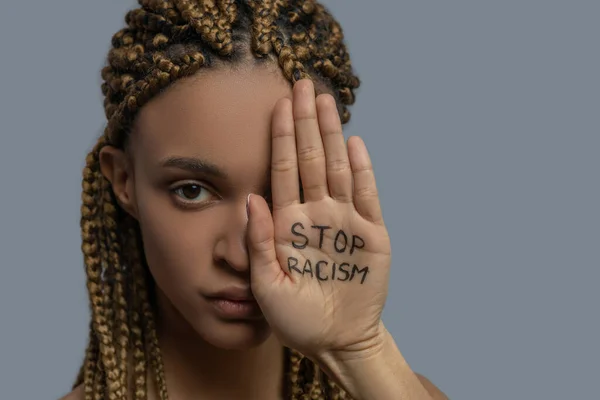 Giovane donna afroamericana che copre metà del suo viso con palma con scritta stop razzismo — Foto Stock