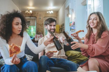 Bir adam ve iki kız kanepede pizza yiyip içki içiyorlar.