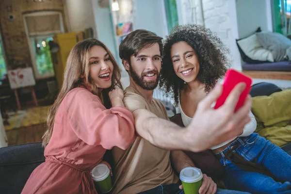 Facet ze smartfonem robi selfie z dwiema dziewczynami w środku. — Zdjęcie stockowe