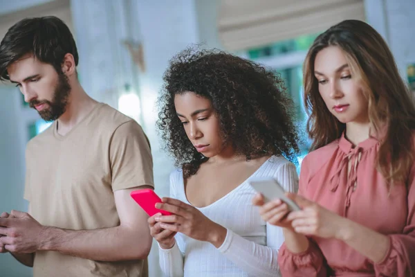 Chico y dos chicas de pie y mirando a los teléfonos inteligentes —  Fotos de Stock