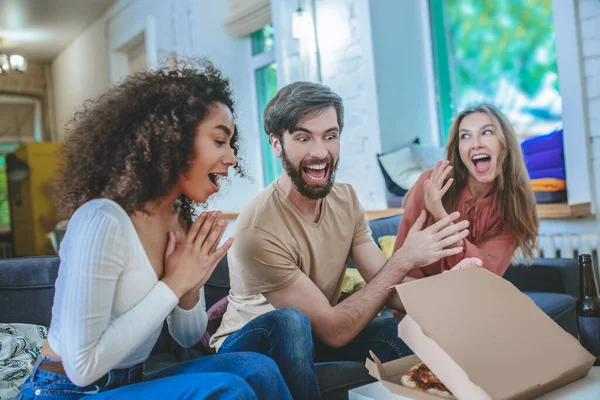 Веселий хлопець відкриває коробку з піцою і двома захопленими дівчатами — стокове фото