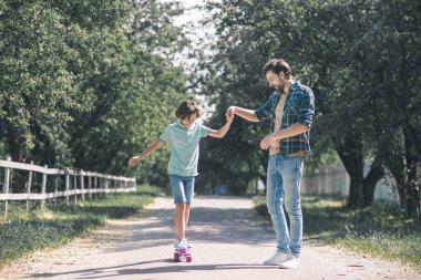 Kaykay süren bir genç, babası elini tutuyor ve öğretmenlik yapıyor.