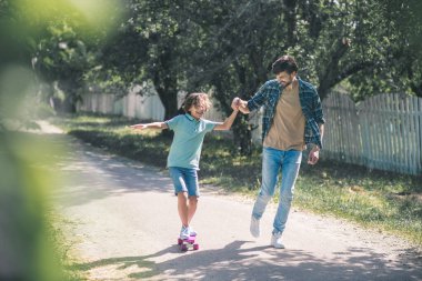 Koyu renk saçlı çocuk kaykaya biniyor, babası onu destekliyor.