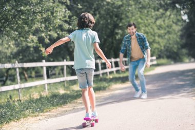Siyah saçlı bir çocuk kaykaya biniyor, babası ona doğru geliyor.