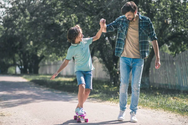 Σκούρα μαλλιά αγόρι ιππασία ένα skateboard, ο μπαμπάς του κρατώντας το χέρι του — Φωτογραφία Αρχείου