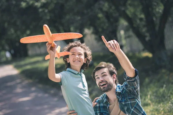 Feliz chico moreno sosteniendo un avión de juguete, su padre señalando al cielo — Foto de Stock