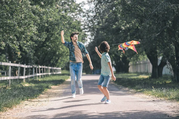 Niño de pelo oscuro y su padre corriendo tras la colorida cometa y luciendo feliz — Foto de Stock